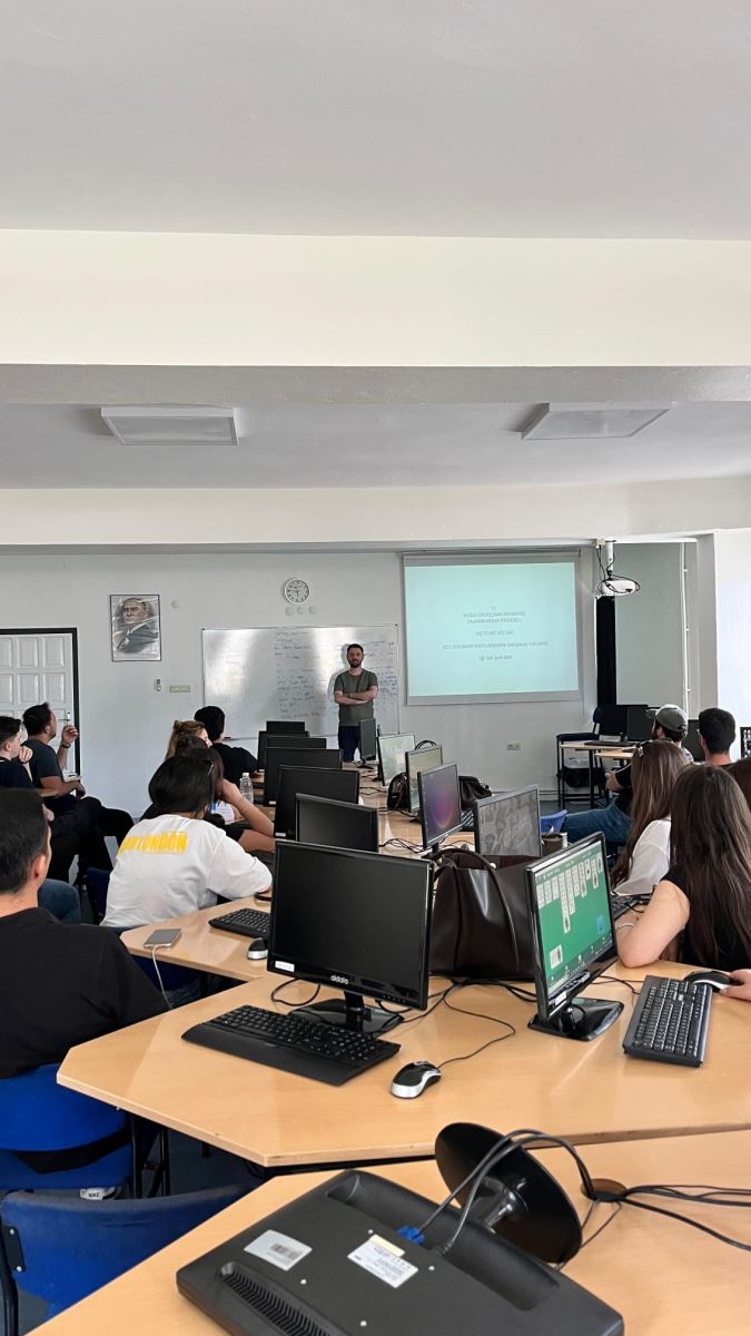 Large windows allow daylight to stream into the room. In the center, there are large light-colored worktables with black computer monitors, keyboards, and cables placed on them. Computer towers can be seen underneath the desks.  Around fifteen university students are seated in the classroom. The students are arranged in a semicircle and are directing their attention toward the left side of the room. The overall atmosphere is calm, attentive, and focused on the lesson.